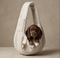 Load image into Gallery viewer, A small brown puppy sits inside a plush cream-colored puppy sling bag with a strap and gold-tone hardware, photographed against a warm beige studio background.