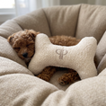 Load image into Gallery viewer, A small brown curly-haired dog rests inside a beige textured pet bed, nestled under a plush bone-shaped cushion embroidered with a monogram logo.
