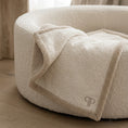 Load image into Gallery viewer, A close-up of a round, plush bouclé pet bed in a warm cream color, with a matching folded throw blanket draped over the front edge. The blanket features a subtle embroidered  PP monogram logo.
