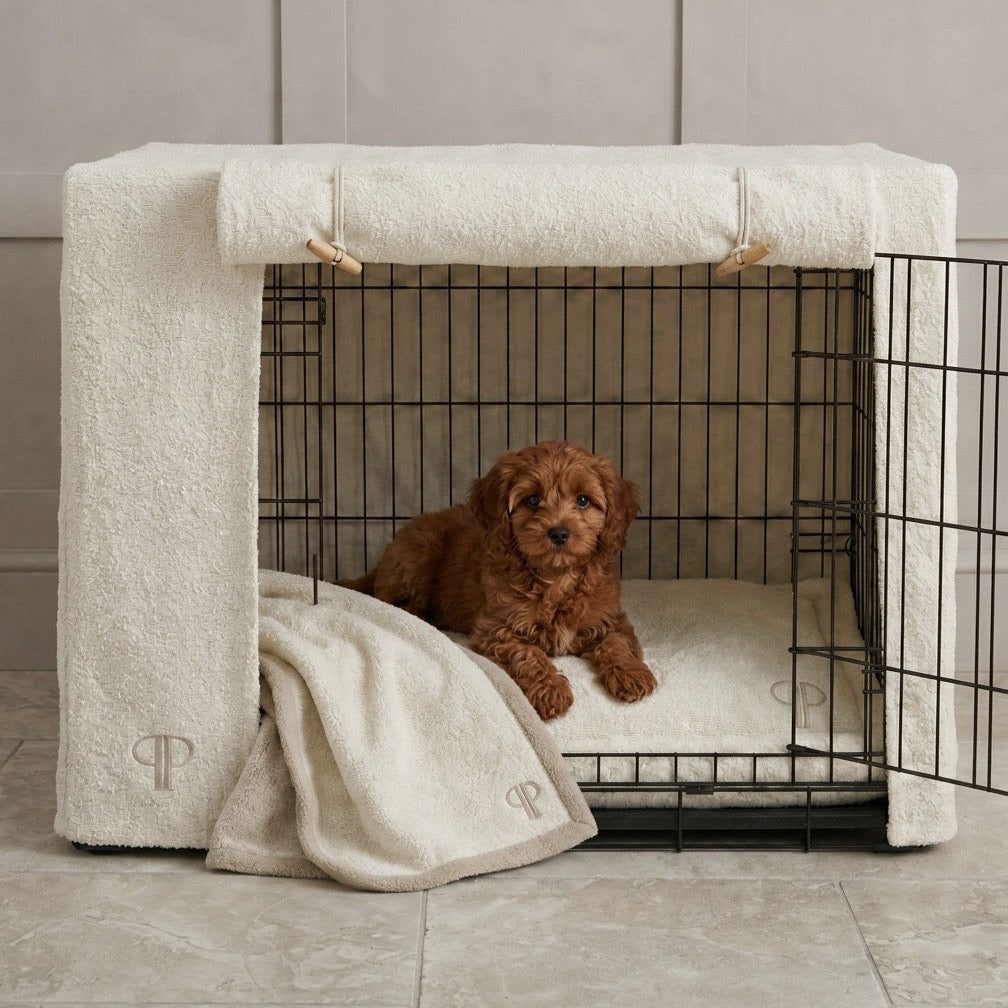 A small red cavapoo puppy sits inside a black wire dog crate covered with cream-coloured Boucle cover, boucle mattress and matching boucle blanket, set against a neutral tiled wall and floor.